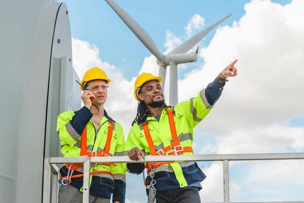 A group of diverse workers in hard hats standing in front of a wind turbine, smiling and looking at the camera. The sky is clear and blue, and the turbine is towering behind them.