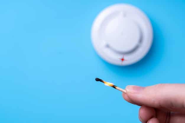 A hand pressing the test button on a smart smoke detector mounted on a ceiling, with a clear view of the LED indicator and the surrounding ceiling area.